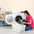 Mosógépbe bepakoló hölgy a mosógép mellett elhelyezett nicol fürdőszobai polccal és szennyestartóval