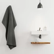 Minimal design fürdőszoba fali mosdóval alatta elhelyezett polccal sötétszürke fürdőlepedővel és a modsón elhelyezett szappanadagolóval