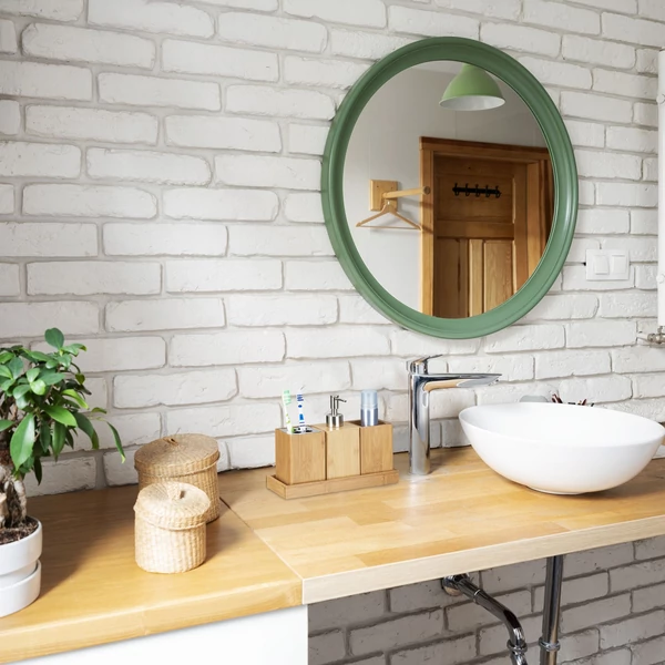 Nagyméretű kerek zöld tükör és rakott fehérre festett téglafal a fürdőszobában  kerámia mosdó mellett elhelyezett bambusz szappanadagoló fogkefe és fogkrém tartó