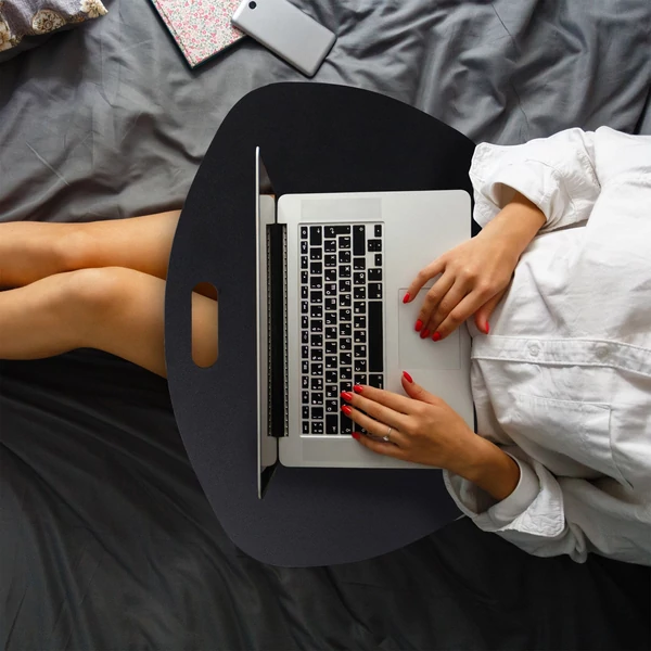 Az ágyból dolgozó fiatal hölgy az ölében elhelyezett fekete színű laptoptartó párnáján elhelyezett notebookjával