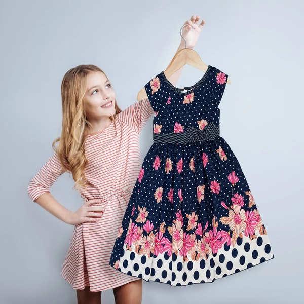 Rózsaszín ruhás kislány a kezében tartott fa gyerek vállfán elhelyezett fekete színes pöttyös ruhájával