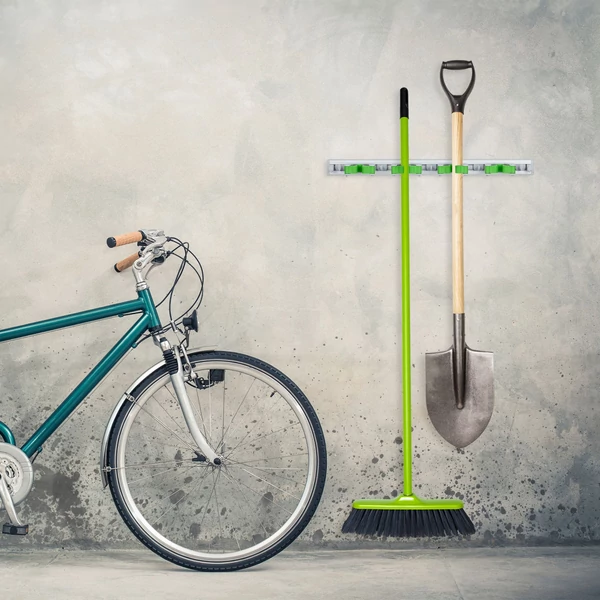 A garács vakolatlan betonfalára felszerelt egy bicikli felett elhelyezett kerti eszközök tárolására alkalmas tartó ásóvl és egy seprővel