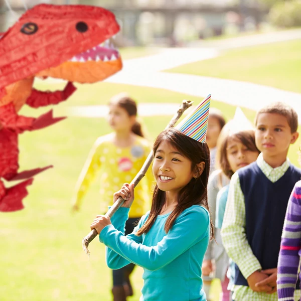 Kisebb gyerekcsoport közepén egy kislánnyal ahogy éppen szétveri a piros dinó pinatat