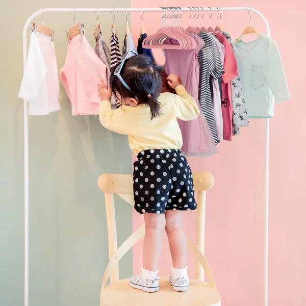 Kislány éppen egy ruhaállványt pakol a ruhaáival amik rózsaszín bársony gyerek vállfákon vannak