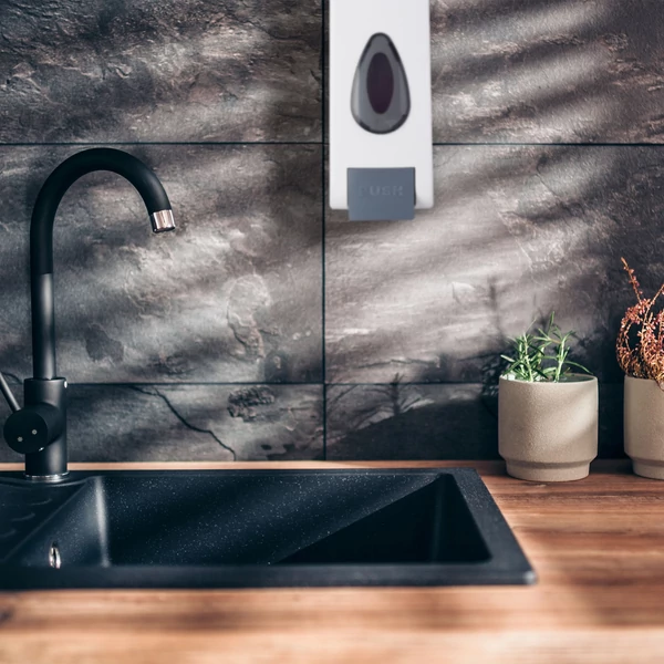 Fehér színű fali mosogatószer adagoló szürke csempefalon a fekete gránit mosogató felett feltöltve kézi mosogatószerrel